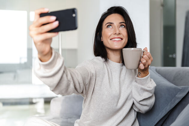 室内30多岁的美女坐在明亮公寓的沙发上用手机自拍的照片生活手机电话
