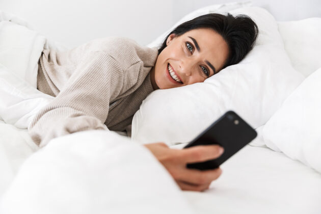 谎言图为30多岁的快乐女人在家里穿着白色亚麻布躺在床上使用智能手机吸引手机公寓