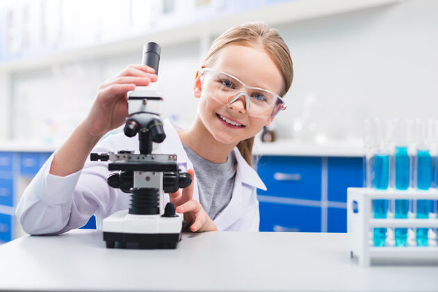 女孩专业的快乐实验室可爱乐观的女孩一边戴着安全眼镜一边触摸显微镜一边看着相机乐趣玻璃器皿实验室