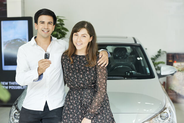 快乐一对年轻夫妇拿着新车钥匙 站在汽车陈列室拥抱微笑的照片客户男人展厅