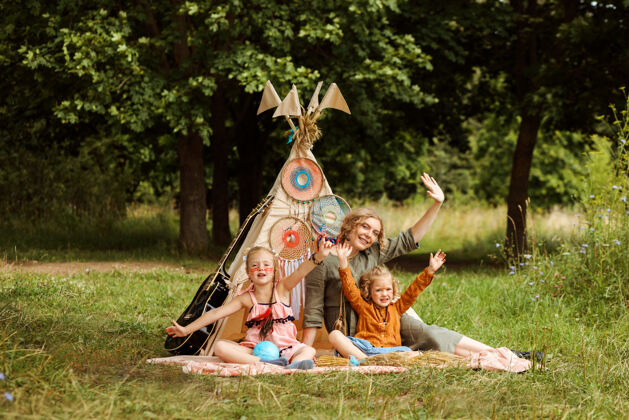 森林妈妈和她的两个女儿坐在公园里的wigwam装饰旁边挥手夏天母亲节户外