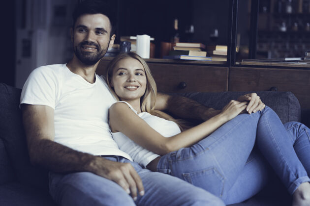 年轻笑容可掬的年轻夫妇在家里放松和看电视男人远程室内