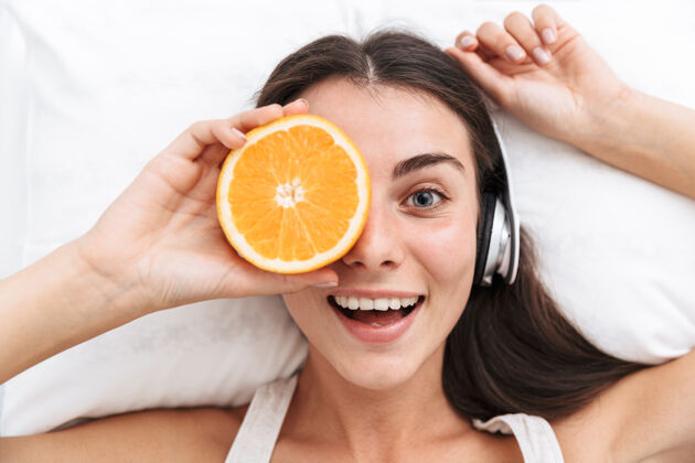 白种人俯视图：一位年轻漂亮的女士躺在床上 手里拿着橘子片 戴着耳机听音乐水果床电子