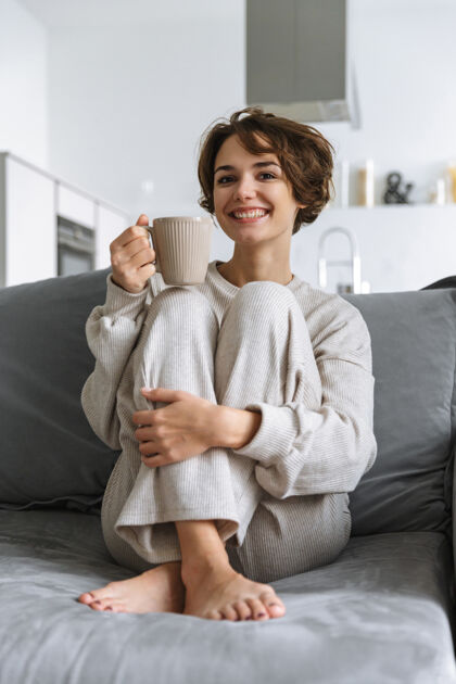 平静快乐的年轻女子坐在家里的沙发上 手里拿着杯子生活茶吸引