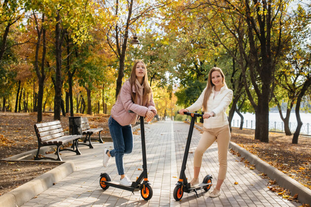 车辆在一个温暖的秋日里 两个年轻漂亮的女孩在公园里骑着电动滑板车周末年轻人步行