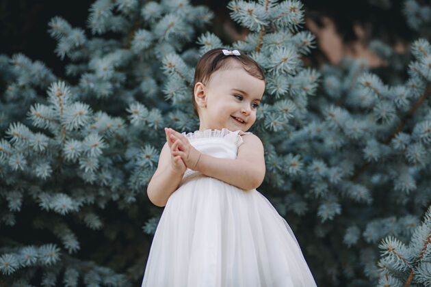小可爱有趣的白裙娃娃在松树前拍手可爱乐趣公园