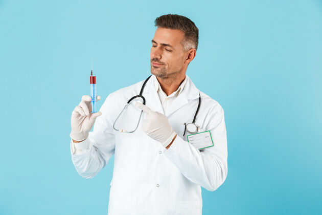 白种人带着听诊器的成熟医生的肖像 戴着手套 拿着注射器 孤立地站在蓝色的墙上肖像手套外套