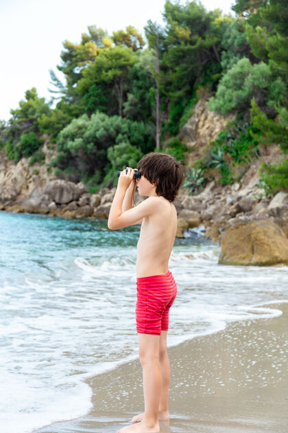青年一个小男孩呆在海边的海滩上 用望远镜看东西探索年轻望远镜