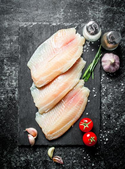 健康把罗非鱼鱼片放在石头板上 用西红柿 蒜瓣和香料放在黑色的乡村桌子上清洁罗非鱼晚餐