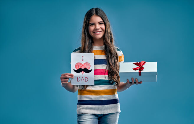 美丽一个穿着条纹多色毛衣的可爱女孩手里拿着一个礼物盒和一张卡片 送给她爸爸 一张蓝色的卡片父亲微笑孩子