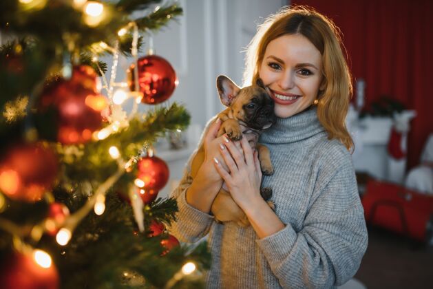 冬天美丽的年轻女子带着一只小狗在圣诞庆典的背景下室内拥抱圣诞树