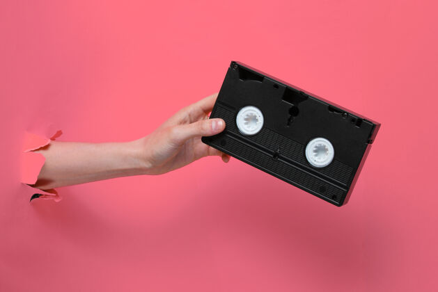 塑料女性手拿着盒式录像带穿过撕破的粉色纸背景：极简主义复古概念磁带手臂模拟