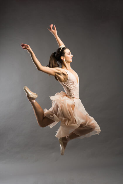 动作年轻漂亮的芭蕾舞演员在浅灰色的工作室背景上穿着米色泳装在尖头上摆姿势颜色拖鞋跳跃