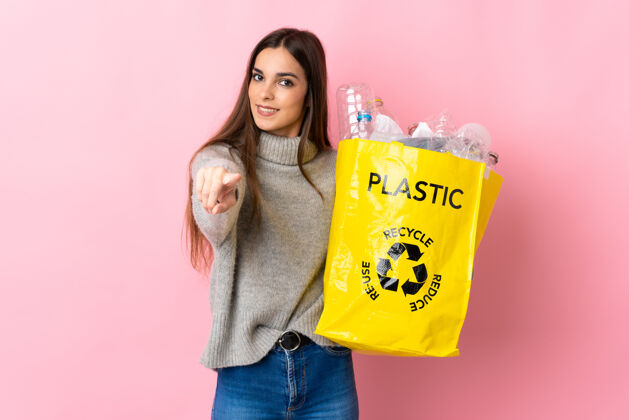 塑料年轻的白种女人拿着一个装满塑料瓶的袋子 在粉红色的背景上用快乐的表情指着前面环境女孩回收