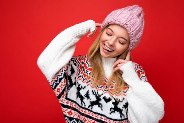 人照片上的美丽幸福有趣快乐的年轻金发女子隔着红墙 穿着冬天的毛衣 戴着粉色的帽子 低头看着 玩得很开心魅力时尚年轻