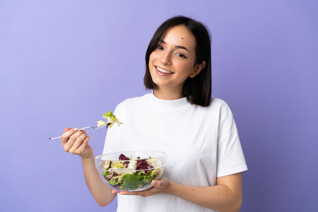 饮食被隔离在紫色墙壁上的年轻白种女人拿着一碗沙拉 表情愉快年轻蔬菜素食