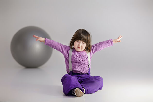 脸小可爱的女孩 穿着紫色衣服 双臂张开 灰色背景上有一个健身用的大球女孩微笑保健
