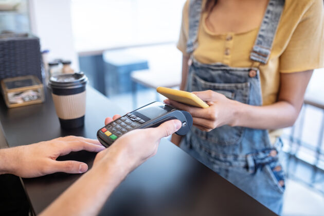 两个人付款 订单号：女纤细的手拿着黄色的智能手机靠近咖啡馆柜台的海报咖啡店付款Pos终端