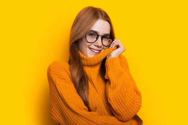 温暖迷人的白种女人 满脸雀斑 红发 戴着眼镜 穿着针织毛衣 在黄色的墙上摆着姿势魅力针织温柔