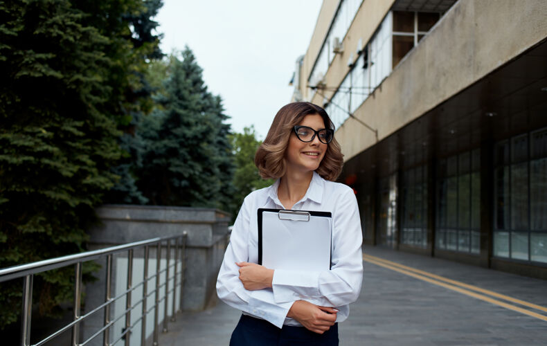 聪明戴着眼镜 穿着白衬衫 手里拿着文件的快乐女商人在街上的大楼附近城市自信女性