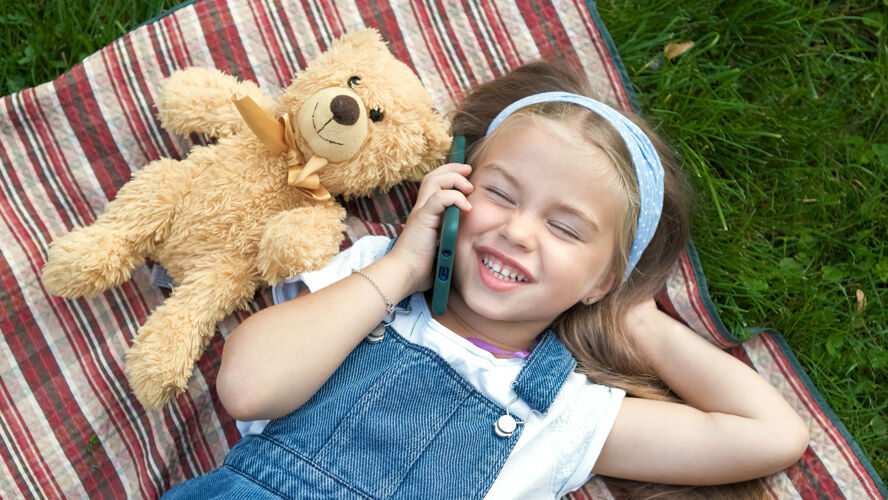 笑夏天 一个快乐的小女孩躺在绿色草坪上的毯子上 泰迪熊在讲手机户外小漂亮