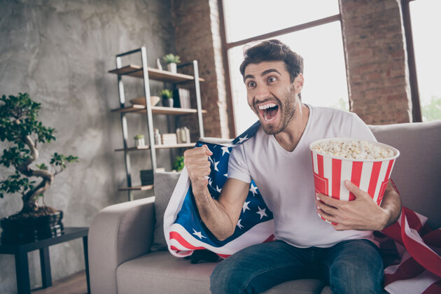 民主照片中混血疯狂的阿拉伯小伙子坐在沙发上拿着爆米花桶举起拳头支持美国足球队比赛肩上挂着美国国旗外套平房室内国旗支持庆祝