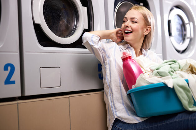清洁开朗的女人开心地坐在那里等着洗完的衣服 微笑着 手里拿着一盆脏衣服和粉红色的洗衣粉瓶家庭主妇洗衣服微笑