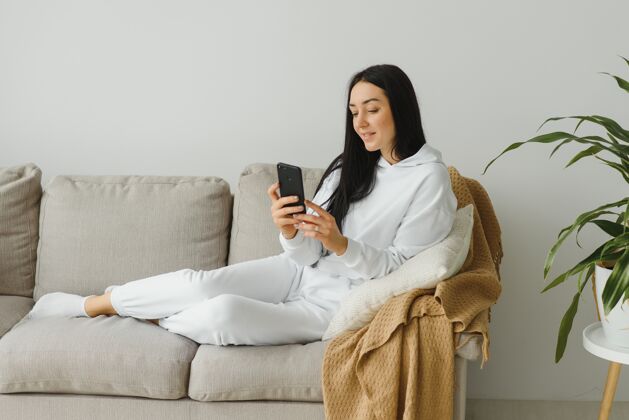 坐着一个快乐的年轻女子在家里的沙发上休息时使用手机的画像年轻快乐美丽