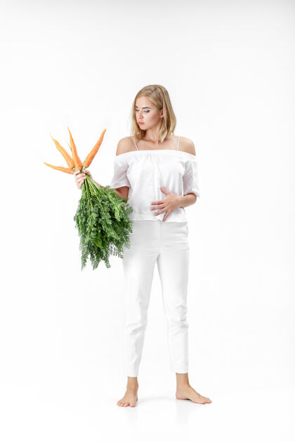 生一个穿着白衬衫的金发美女 手里拿着一根新鲜的胡萝卜 上面有绿叶背景.健康还有节食背景橙色脸