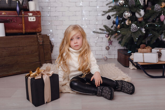 衣服一个时髦的小女孩在圣诞树旁摆姿势 期待圣诞节的到来地板木头房间