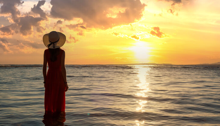 剪影穿着红色长裙 头戴草帽的年轻女子站在海边的海水中 欣赏着初夏的朝阳日出海滩新鲜