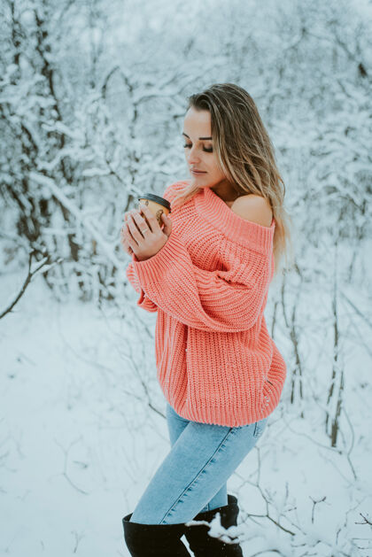 雪花一个穿着粉色宽松毛衣和牛仔裤 手里拿着咖啡的漂亮女孩 在寒冷的雪域森林里年轻享受牛仔裤