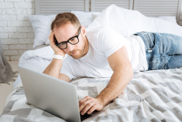 当代做我的自由职业者工作魅力运动迷人的男子躺在床上 使用笔记本电脑 同时表达兴趣和上网内部信息积极性