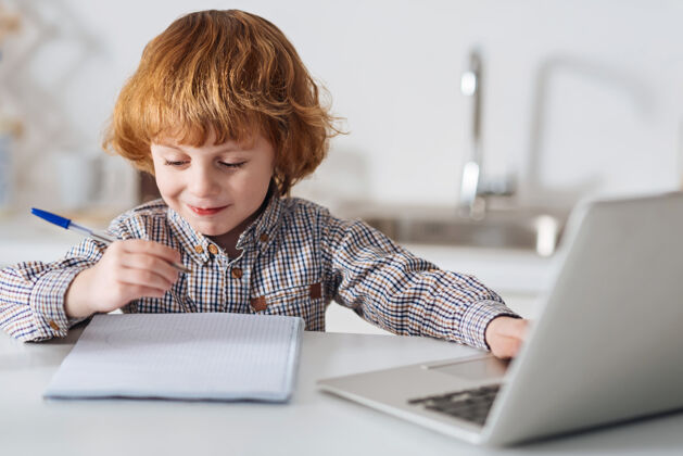 笔记本电脑才华横溢的小创意作家勤劳可爱的男孩在网上找东西 坐在厨房的桌子旁写下来发展休闲勤奋