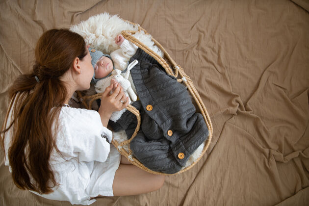 童年妈妈和宝宝 在温暖的毛毯下 戴着温暖的针织帽 甜蜜地睡在柳条摇篮里妈妈幼儿温暖