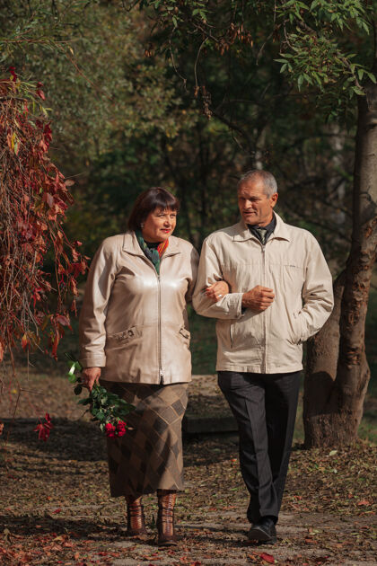 户外一对老夫妇手牵着手 捧着一束红玫瑰走在秋天的公园里60岁森林公园