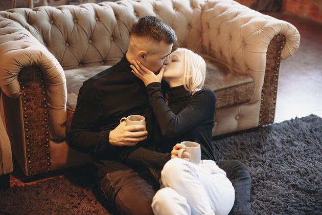 金发一对迷人的年轻夫妇坐在地板上 靠在沙发上 拿着杯子和热咖啡亲吻的画像爱人生活方式