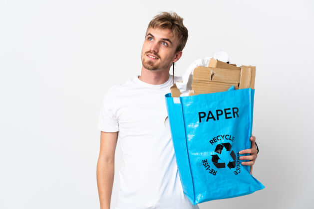 英俊年轻的金发男子拿着一个装满纸的回收袋 在白色的隔离思考着一个想法 同时抬起头来青年垃圾生态