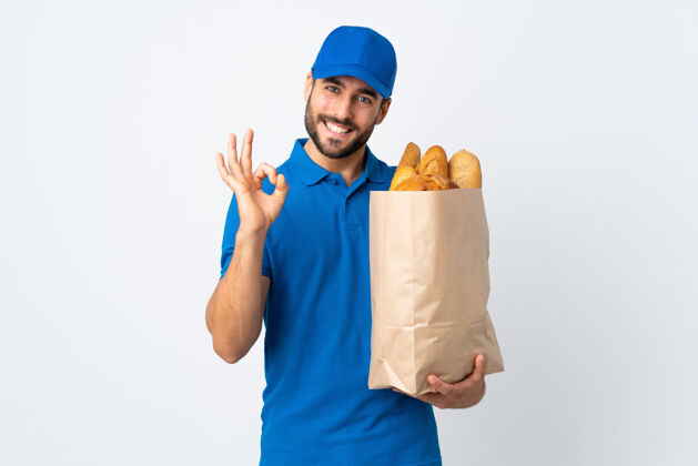 年轻送货员拿着装满面包的袋子 用手指在白色上显示ok标志制服围裙乐观