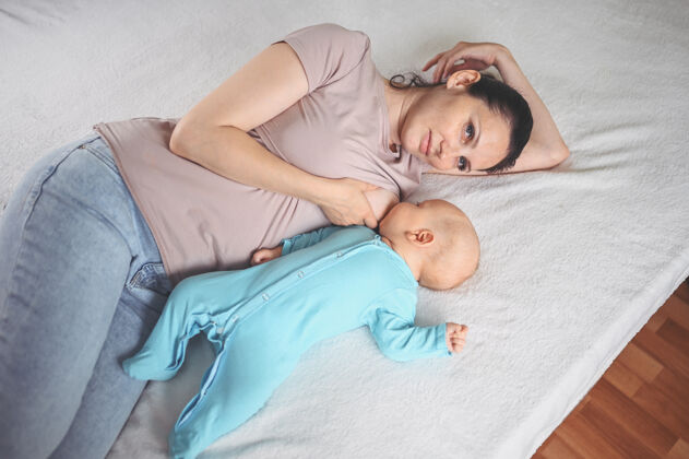 食物年轻的母亲躺在床上 抱着一个穿着蓝色连体衣的可爱的新生婴儿 用母乳给他喂奶婴儿乳房女人