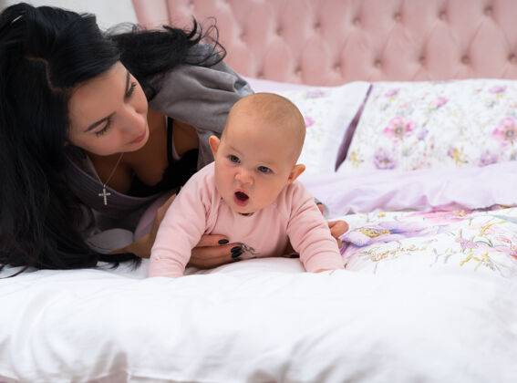 母性年轻的黑发母亲 躺在她的小宝宝旁边 而女孩正试图爬在床上搞笑面已查看从他们的前面复制空间可爱女孩封闭