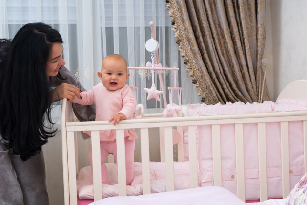 封闭可爱的年轻母亲在托儿所里帮助她微笑的小女儿站在婴儿床里笑室内平衡