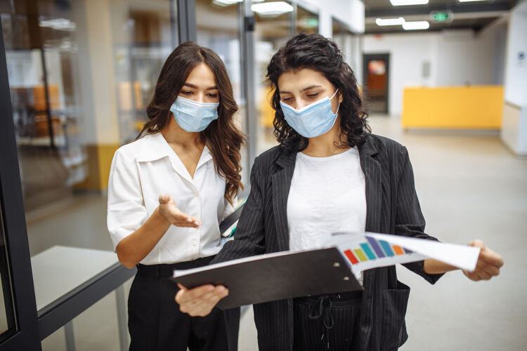 项目两位女商人穿着医用无菌衣在办公室讨论工作问题面具工作在冠状病毒大流行隔离期间笔记本电脑同事文件