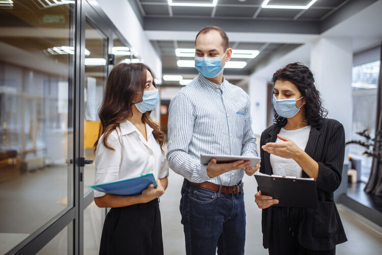 穿着三个戴着医用口罩的同事在冠状病毒大流行隔离期间在办公室走廊讨论业务成长办公室防护