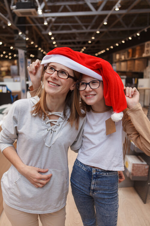 购买垂直拍摄的快乐的母亲和女儿一起戴着大圣诞老人的帽子家具女学生单亲