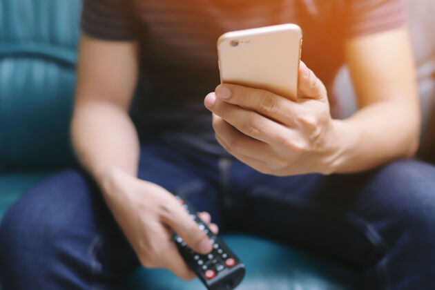 电子年轻人用电视遥控器和移动智能手机坐在椅子上沙发上或者关掉电视遥控器通讯娱乐
