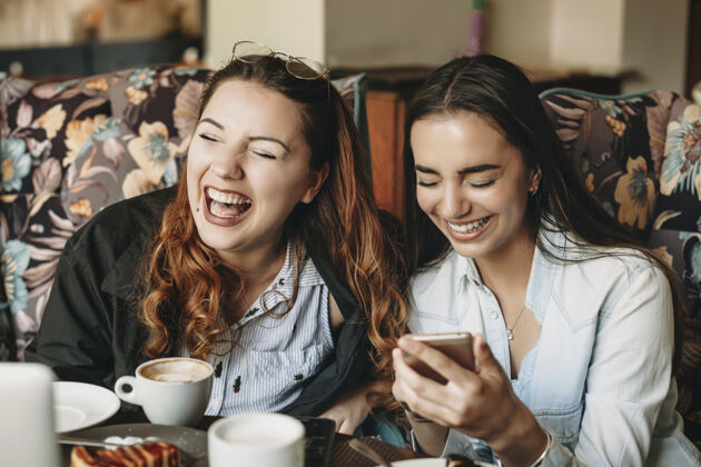 手机两个迷人的女朋友坐在咖啡馆里 闭着眼睛笑着拿着智能手机人乐趣微笑