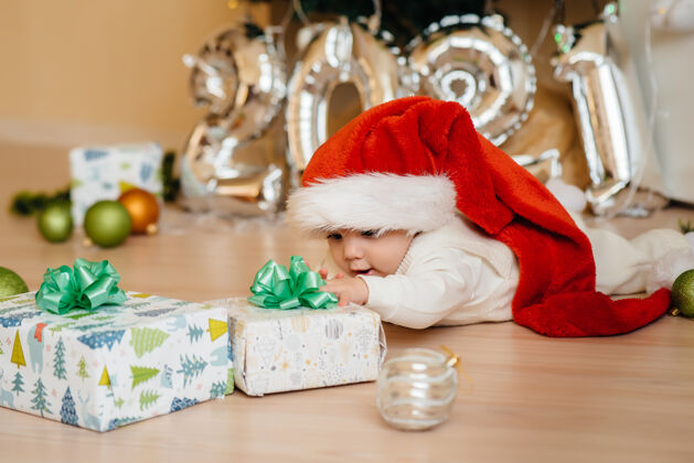 乐趣可爱的微笑的宝宝躺在圣诞树下玩礼物帽子帽子新