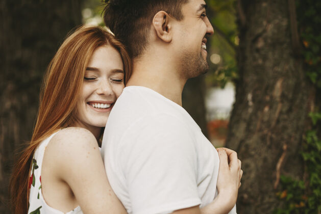 头发迷人的红发雀斑女人闭着眼睛笑着 从背后拥抱她的男朋友 在城市的户外笑着阳光爱成人
