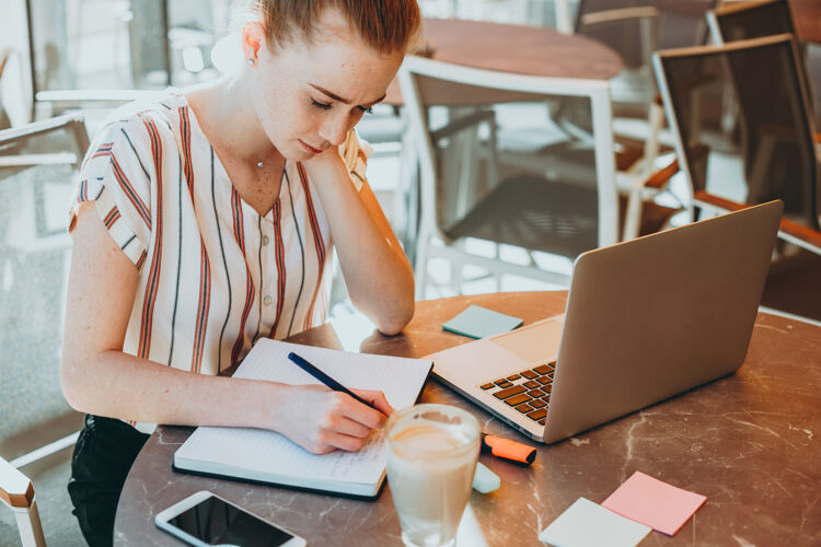社交媒体上图是一位年轻的商务女性 红发 长着雀斑 在咖啡馆的户外笔记本上做笔记 桌上放着一台笔记本电脑笔记在线网络